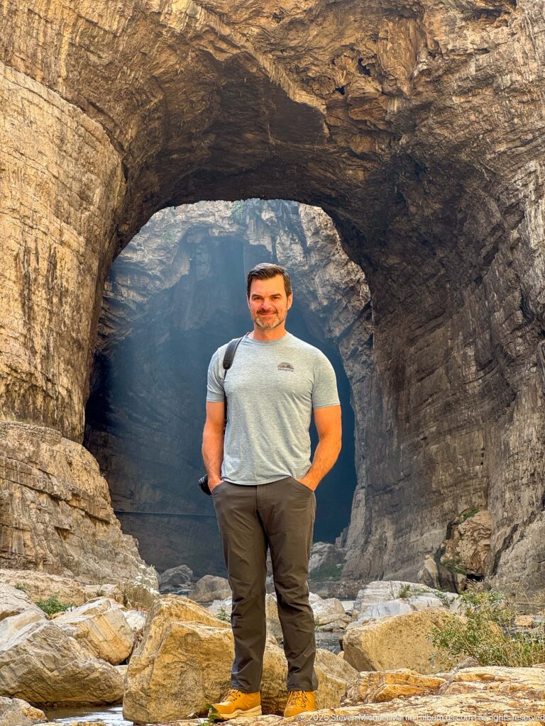 Man standing centered beneath a broad natural rock arch, framed by warm stone and a darker gorge passage.