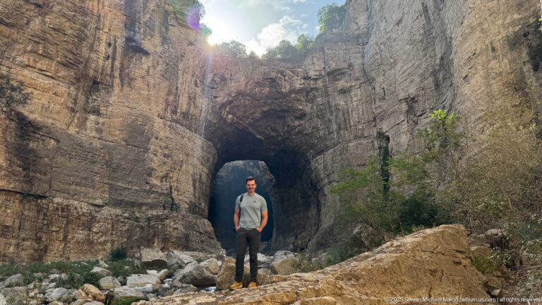 Man standing among boulders before a large dark opening in the layered rock walls of Constantine’s gorge.