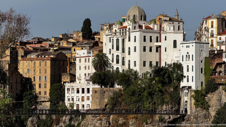 Large white domed building above a narrow pedestrian bridge, surrounded by hillside buildings in Constantine.