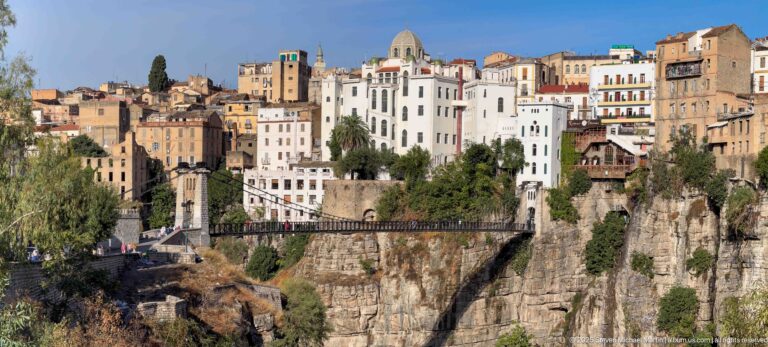 Metal pedestrian bridge spanning a rocky cut below a large white domed building and dense hillside housing in Constantine.
