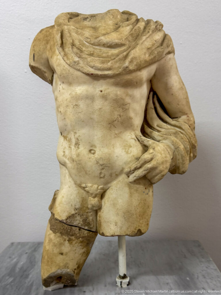 Fragmentary marble male torso with exposed chest and drapery gathered over one shoulder and arm.
