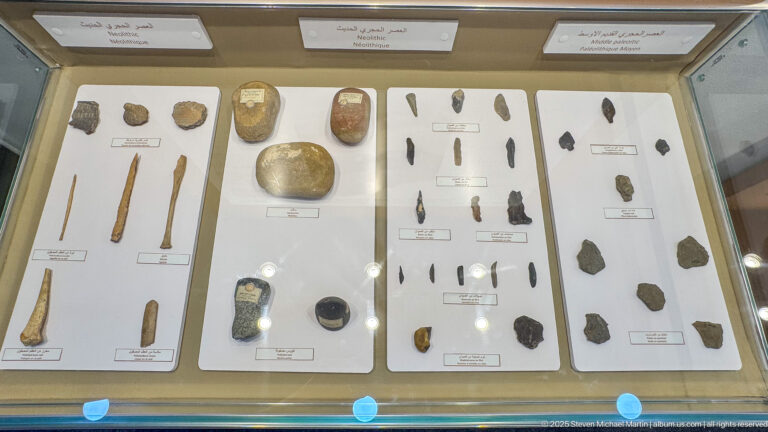 Display case with stone flakes, pointed tools, bone fragments, and labeled prehistoric artifacts arranged on white boards.