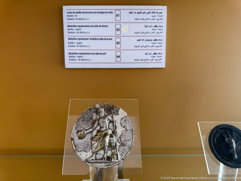 Small circular relief medallion on a clear stand, showing a raised standing figure and surrounding details.