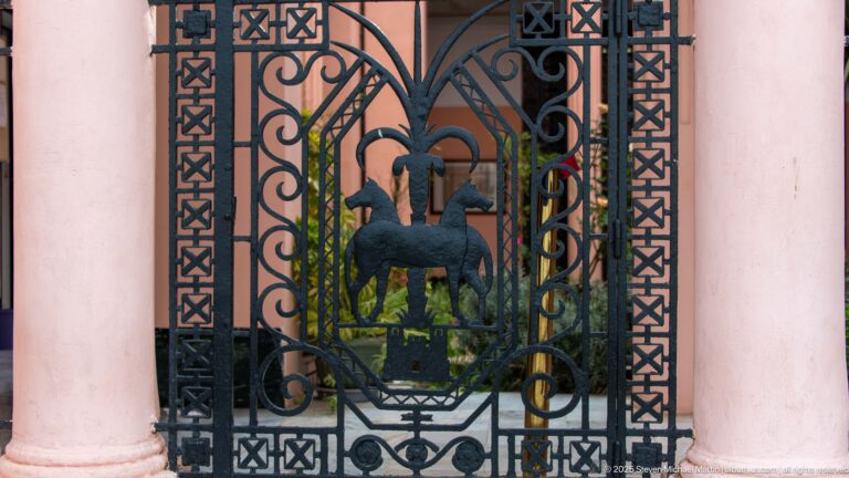 Decorative black iron gate panel between pink columns, with scrollwork and a stylized animal motif.