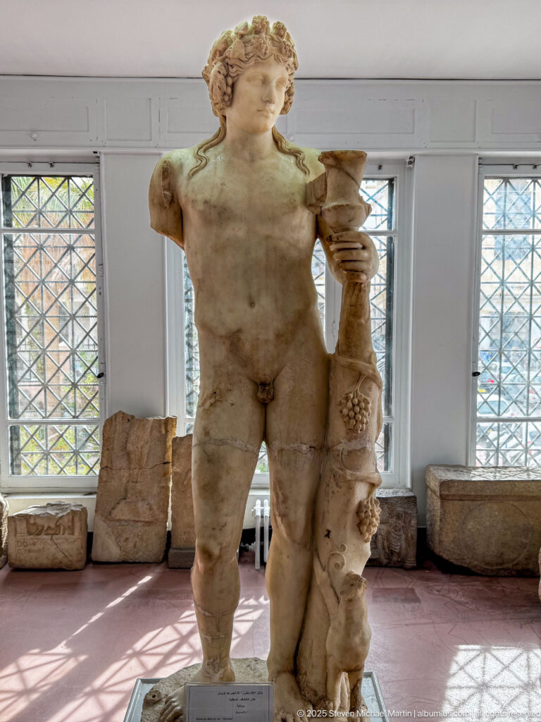 Marble statue of a nude youthful man standing beside a tall fruit- and foliage-decorated support.