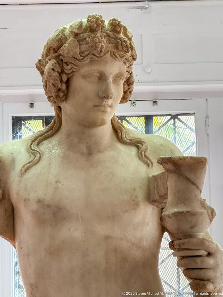 Classical marble youth with long curled hair holding a small cup or vessel in one hand.