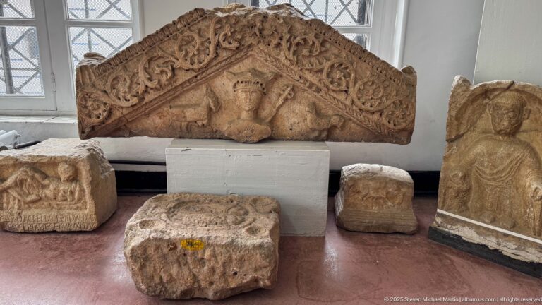 Triangular carved stone relief with a central frontal bust surrounded by dense scrolling vegetal ornament, displayed with smaller fragments.