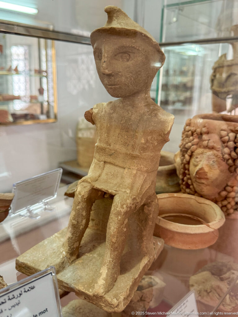 Small seated anthropomorphic clay vessel with rounded head, blocky torso, and two legs, displayed inside a museum case.