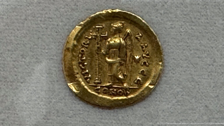 Close-up of a gold coin showing a standing figure surrounded by an inscription and a small mark below.