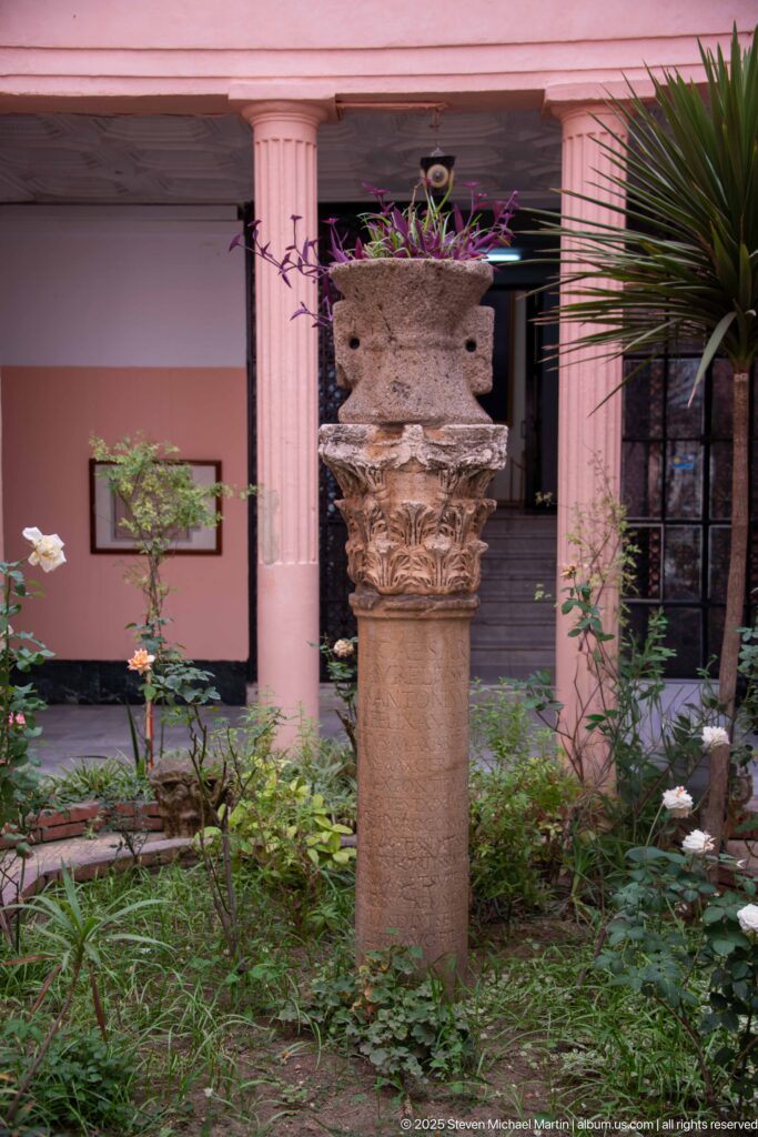 Carved stone capital with leafy ornament standing among plants in the Cirta Museum courtyard.