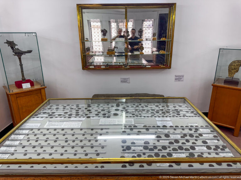 Wide museum-gallery view with a large coin case in front and smaller artifact displays around the room.