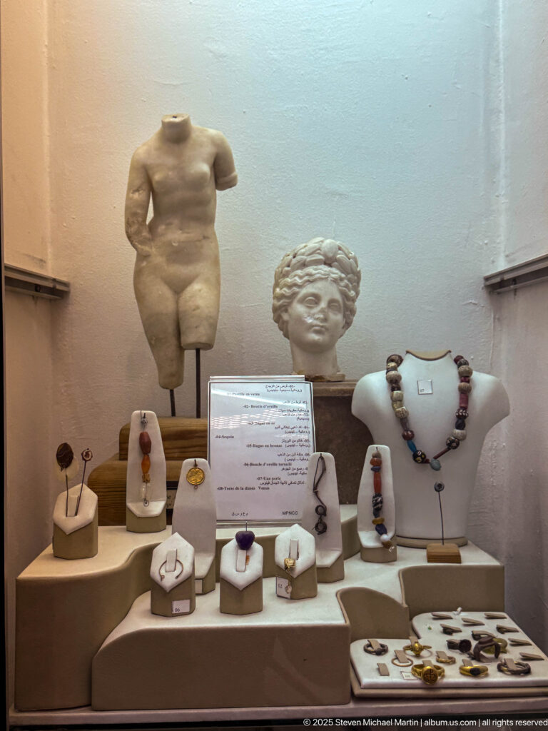 Jewelry and small ornaments displayed on stands before marble torsos and a sculpted head in a museum case.