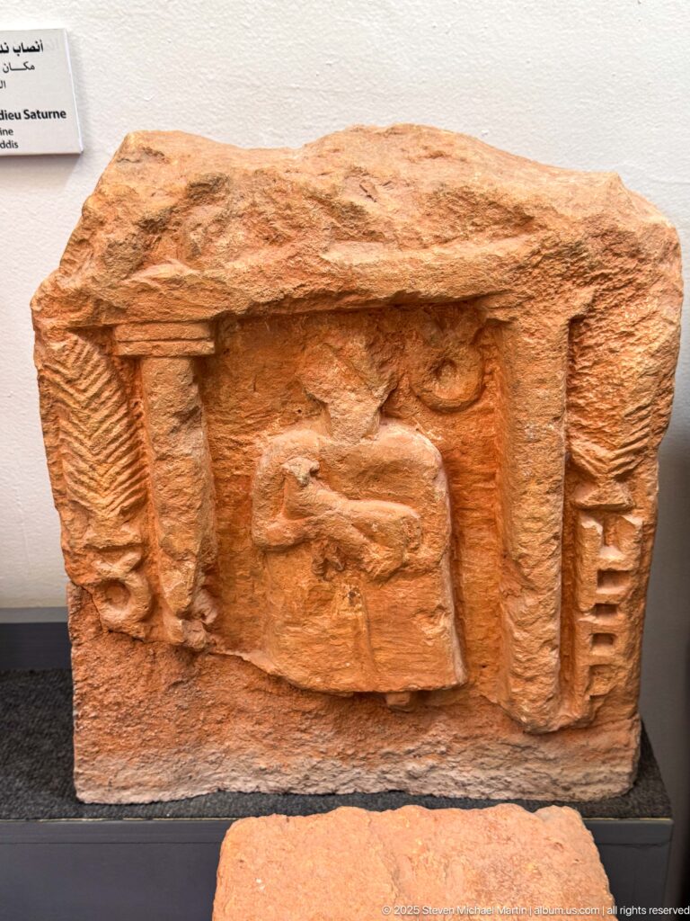 Reddish rectangular relief fragment with a worn central human figure framed by vertical elements beneath a projecting ledge.