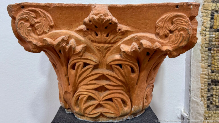 Reddish architectural capital with stylized leaves, curled upper corners, and a symmetrical carved front.