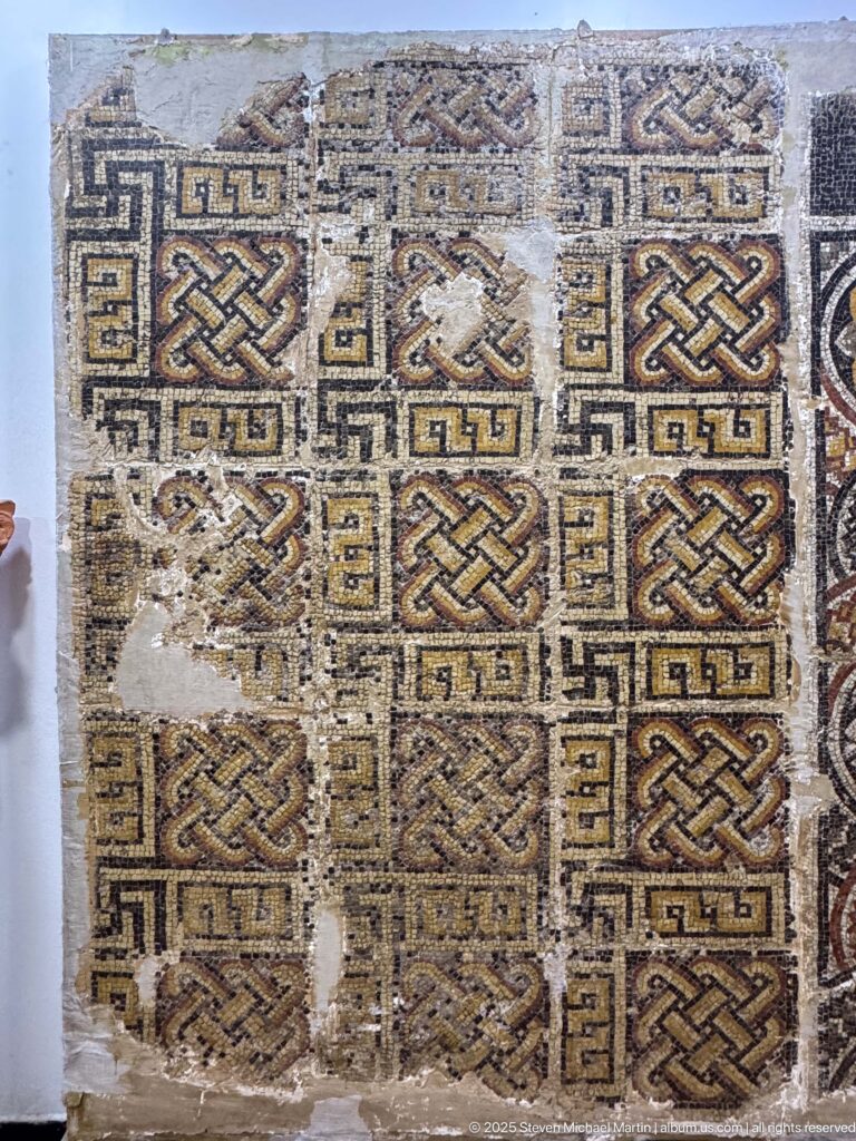 Large geometric mosaic pavement fragment with interlace squares, meander borders, and several damaged or missing areas.