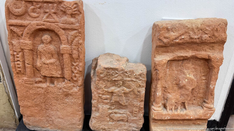 Three reddish carved stone stelae, including standing human figures within simple shrine-like frames.