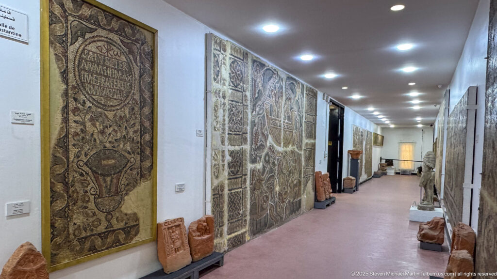 Long museum gallery lined with large mosaics and carved stone fragments under a row of ceiling lights.