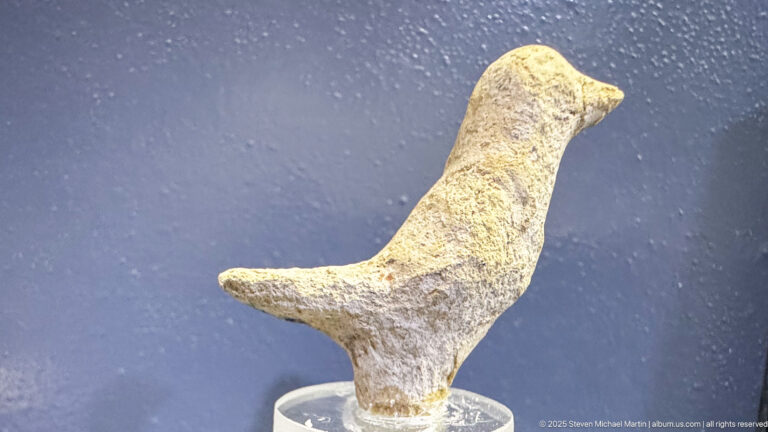 Small pale bird-shaped object with a long neck, pointed beak, rounded body, and short tail.
