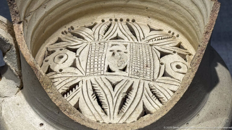 Triangular carved stone panel with a central face, lattice panels, and radiating vegetal ornament.