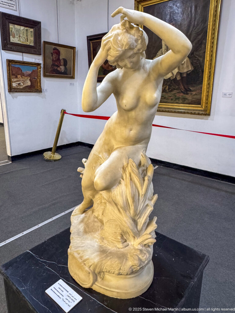 White stone sculpture of a nude female figure seated or kneeling on a rocky base with both arms raised.