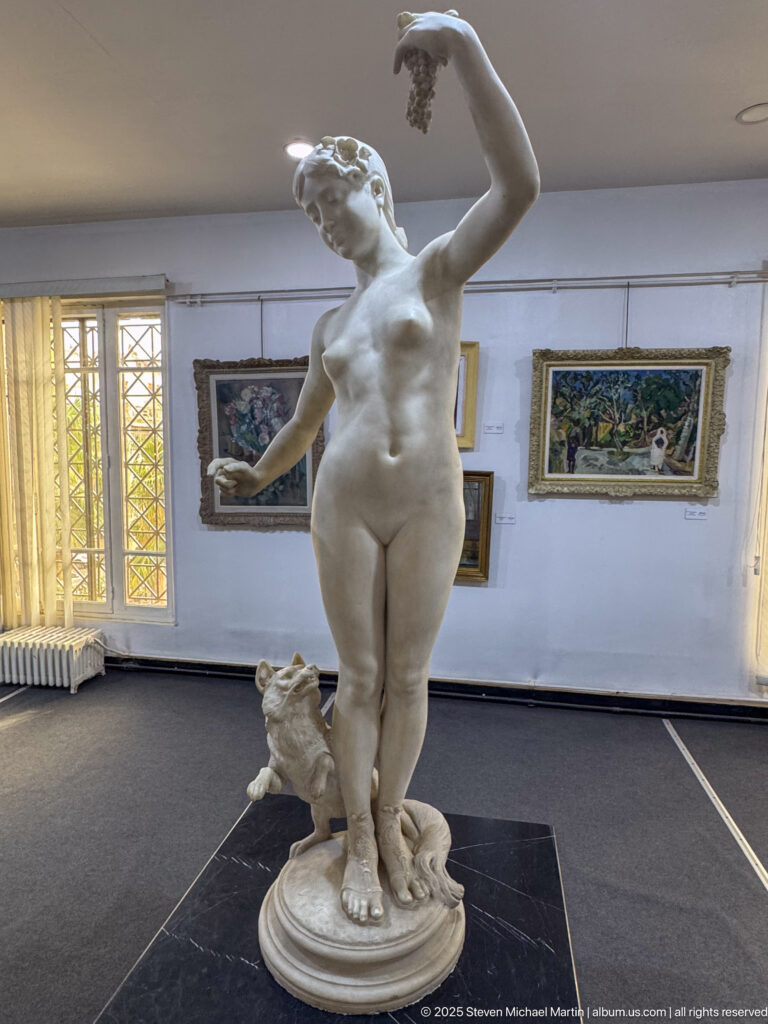 Standing white female nude sculpture with one arm raised and a small sculptural support near her leg.