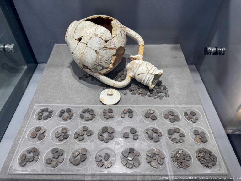 Broken ceramic vessel displayed above many grouped ancient coins in a museum case.
