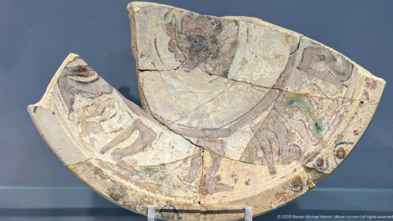 Large curved ceramic fragment painted with partial human figures and geometric bands in red, black, green, and cream.