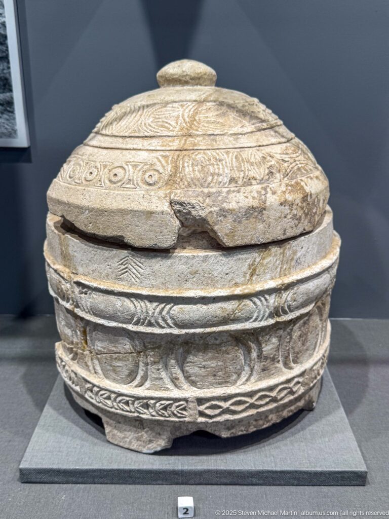 Oval carved stone container with domed lid, scrolling bands, figural relief decoration, and visible breaks along the rim.