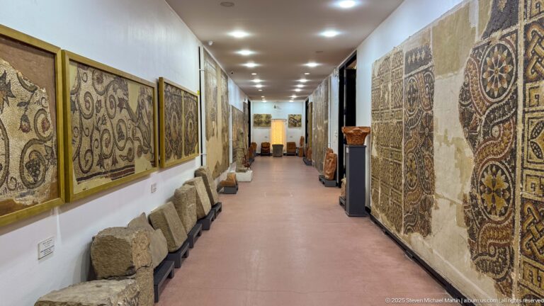 Long gallery corridor lined with large wall-mounted mosaics and stone fragments on low supports.