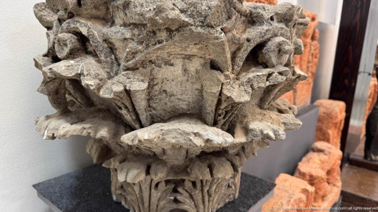 Weathered carved stone capital with layered acanthus leaves, projecting volutes, and broken edges.