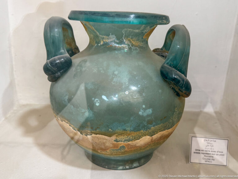 Blue-green ancient glass jar with rounded body, broad rim, and two thick loop handles.