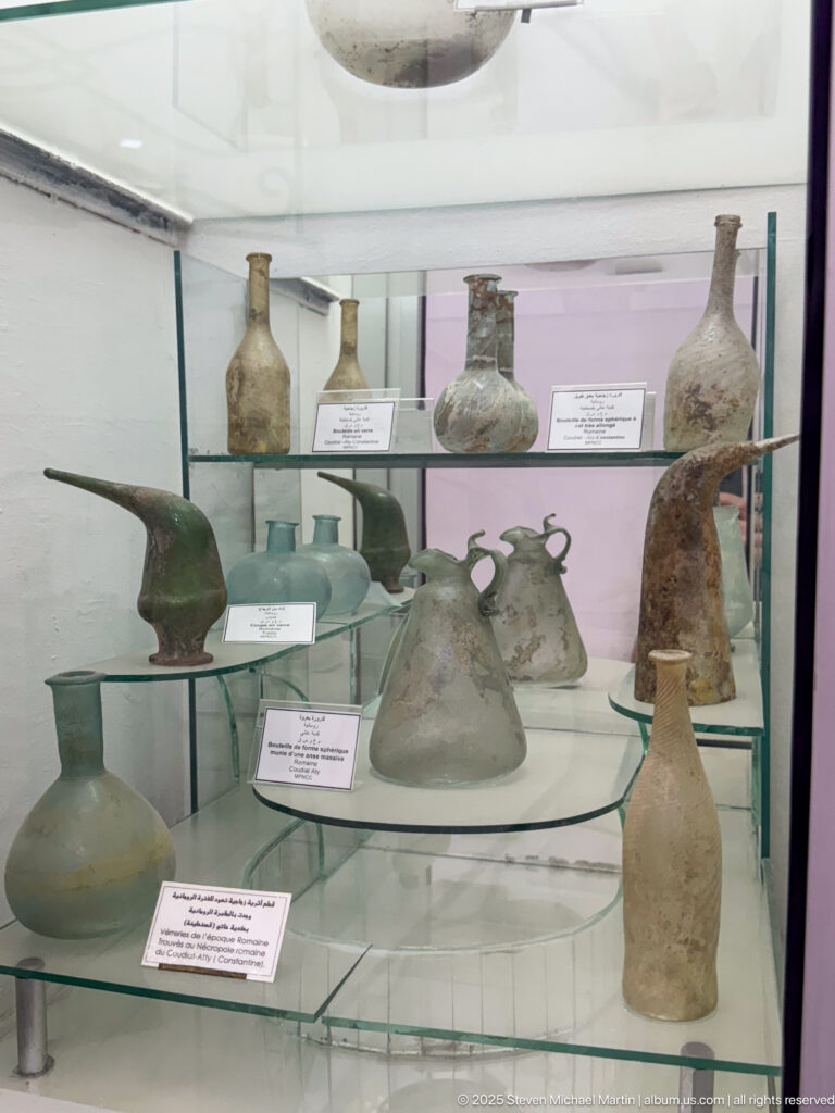 Display case filled with ancient glass bottles, flasks, jugs, and handled vessels on glass shelves.