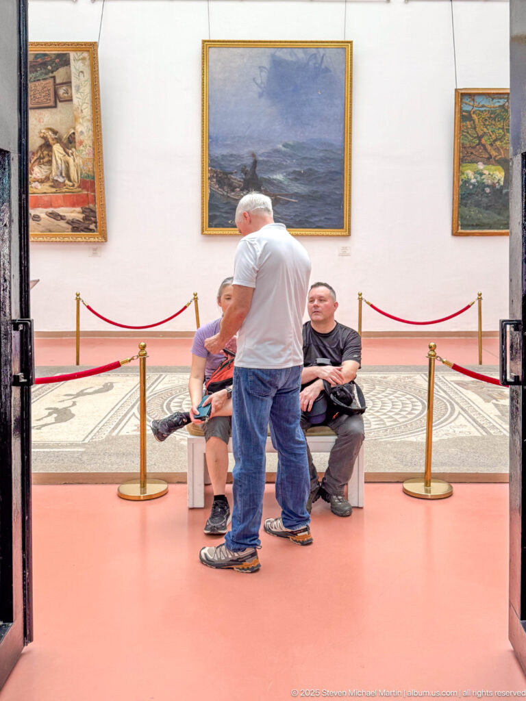 Visitors seated and standing in a museum gallery with framed paintings and a roped-off mosaic floor display.
