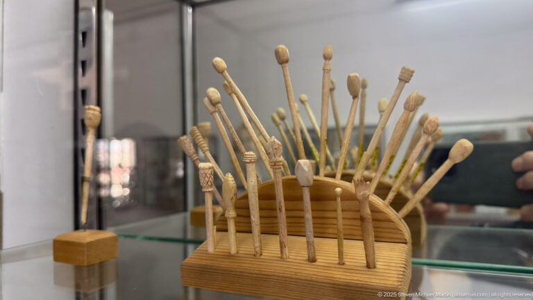 Display of slender pale carved pins and small implements with varied decorative tips in a wooden holder.