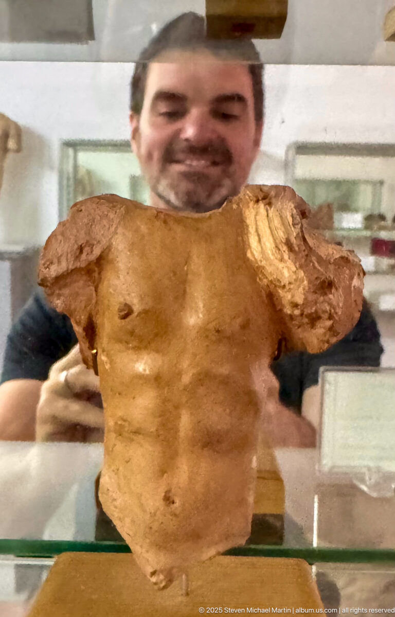 Fragmentary male torso sculpture with missing head and arms, shown behind reflective museum glass.