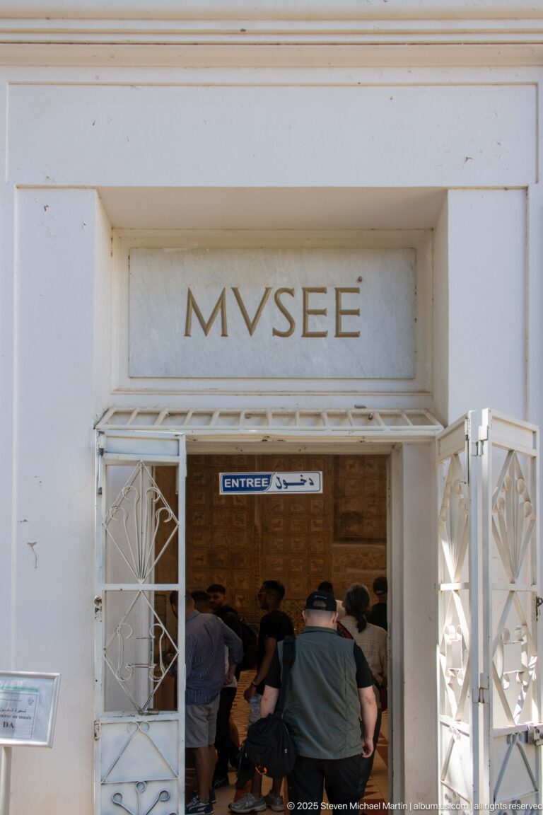 Visitors entering Djemila Museum beneath the carved word MVSEE, with white metal gates open on both sides.