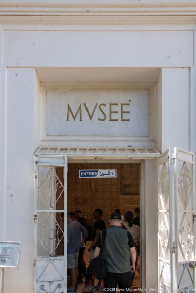 Visitors entering Djemila Museum beneath the carved word MVSEE, with white metal gates open on both sides.