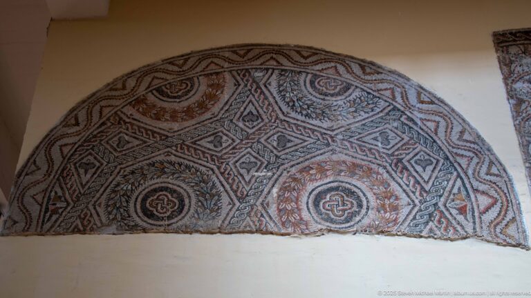 Semicircular Roman mosaic divided into geometric compartments with rosettes, interlace, and red, black, and cream tesserae.