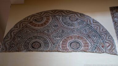 Semicircular Roman mosaic divided into geometric compartments with rosettes, interlace, and red, black, and cream tesserae.