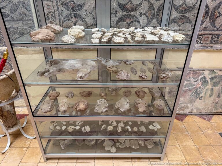 Glass museum case with small pottery and carved archaeological fragments arranged on shelves, with Roman mosaics behind it.