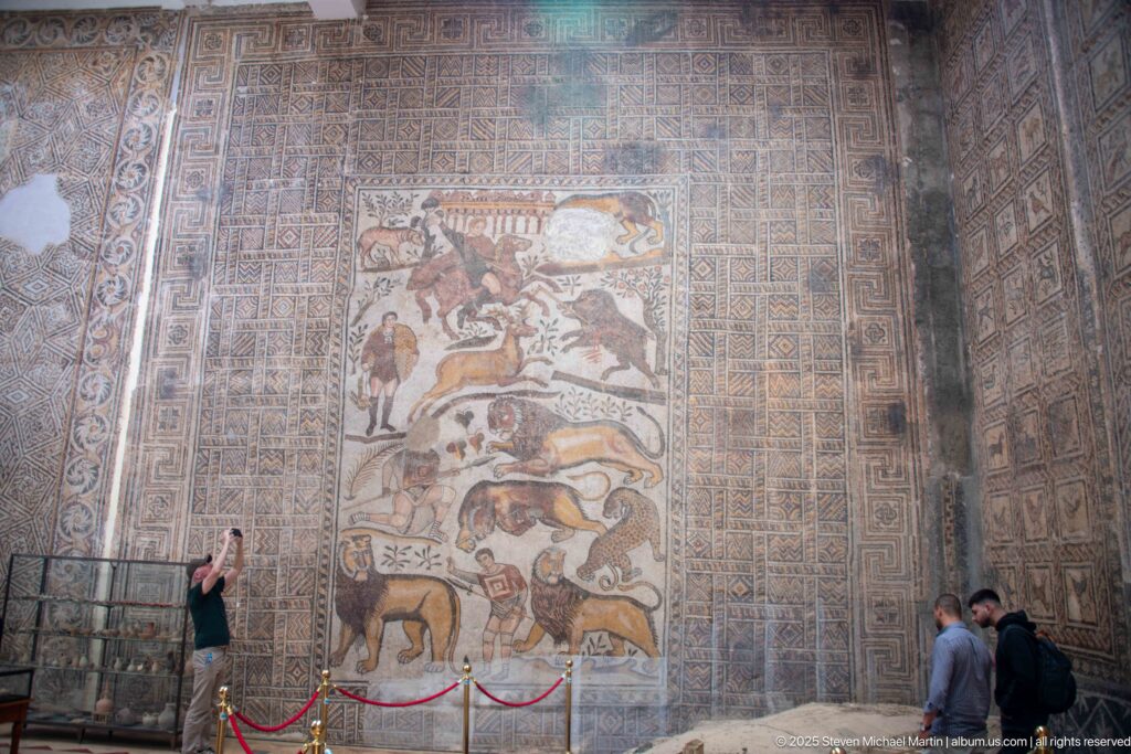 Great Hunt Mosaic from the House of Bacchus mounted on a tall museum wall, with visitors standing below.