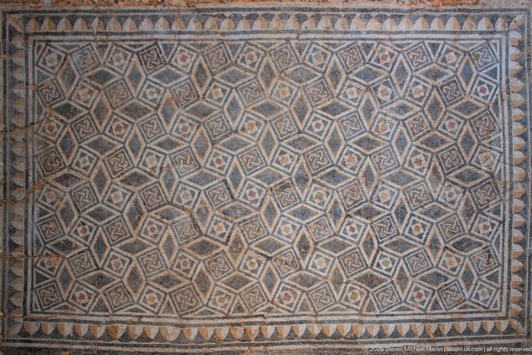 Roman mosaic filled with a dense diamond grid and small motifs, edged by a row of triangular geometric forms.