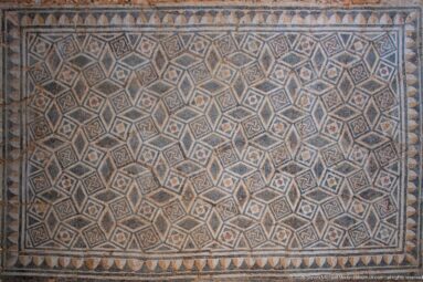 Roman mosaic filled with a dense diamond grid and small motifs, edged by a row of triangular geometric forms.