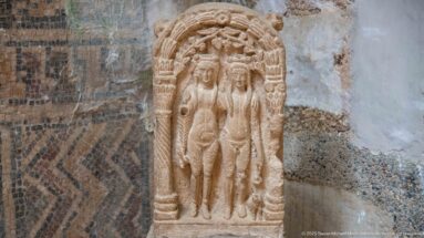 Stone relief of the Three Graces, three nude women standing together beneath a rounded carved frame.