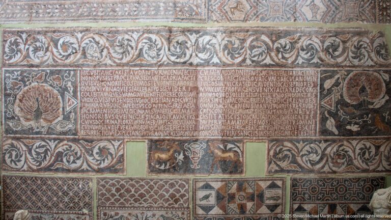 Roman mosaic with a central Latin inscription surrounded by scrolling foliage, animal motifs, vessels, and geometric borders.