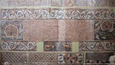 Roman mosaic with a central Latin inscription surrounded by scrolling foliage, animal motifs, vessels, and geometric borders.