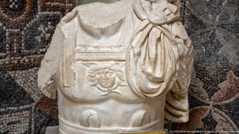 Fragmentary Roman marble torso wearing a decorated cuirass and draped garment, displayed against a mosaic.