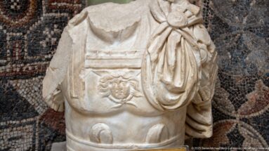 Fragmentary Roman marble torso wearing a decorated cuirass and draped garment, displayed against a mosaic.