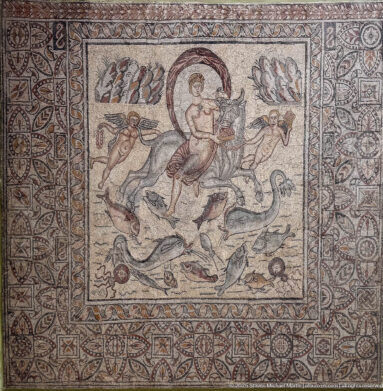 Figurative Roman mosaic with a male rider or hunter surrounded by a lion and other wild animals within ornate borders.