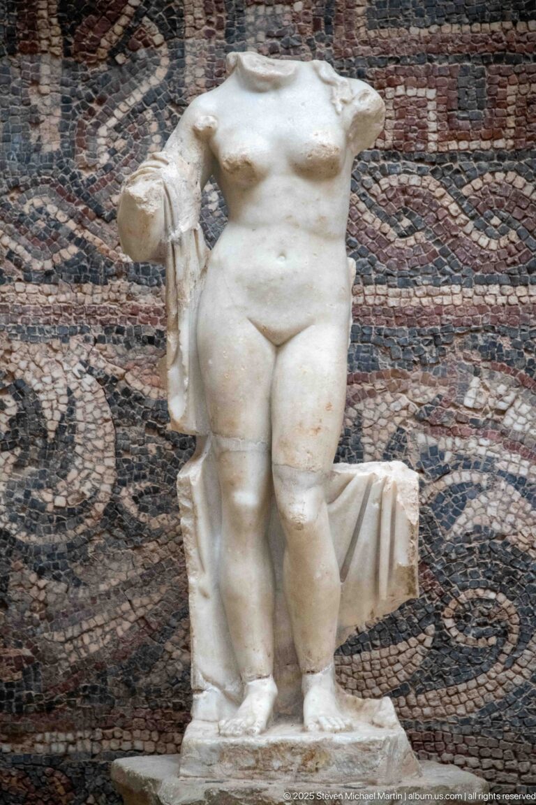 Headless Roman marble female nude with side drapery and support, displayed before a patterned mosaic.